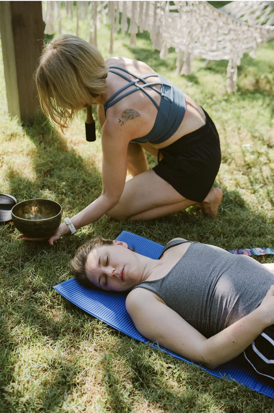 One person resting on a mat outdoors while another places a singing bowl on their chest