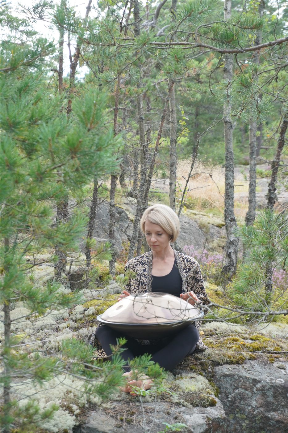 Natasha sitting on a rock in a pine forest with a handpan resting on her lap