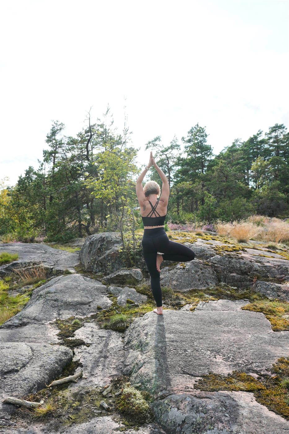 A person standing in tree pose on a rocky outcrop, arms lifted against an open sky