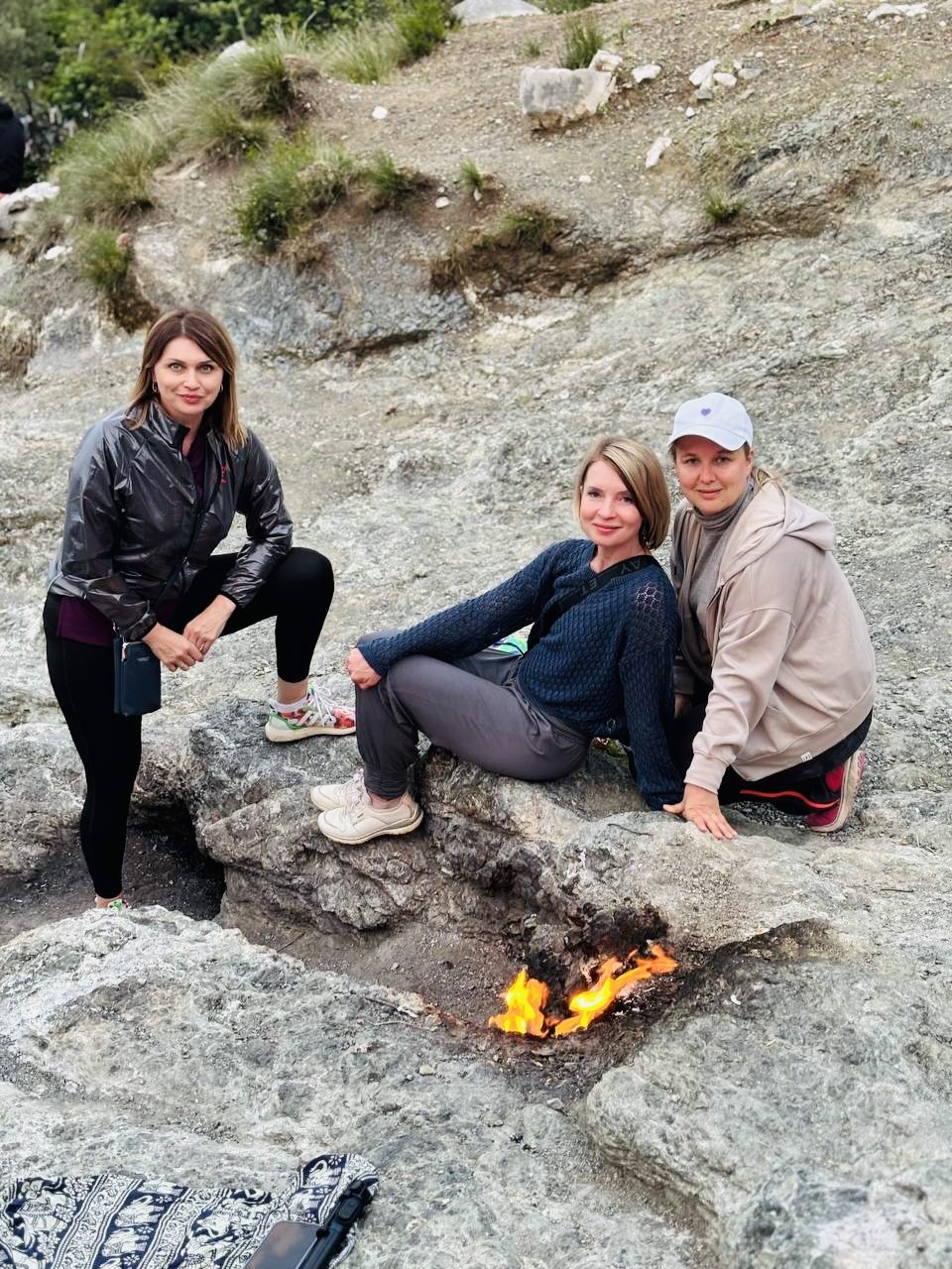 Three people sitting on warm rock beside natural flames rising from the ground at Çıralı