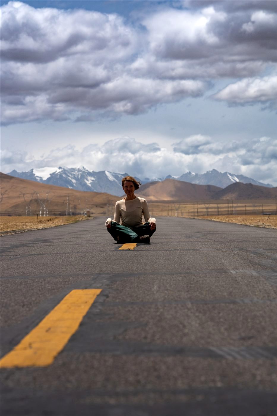 A person sitting cross-legged in the middle of an empty road, mountains ahead under a big sky