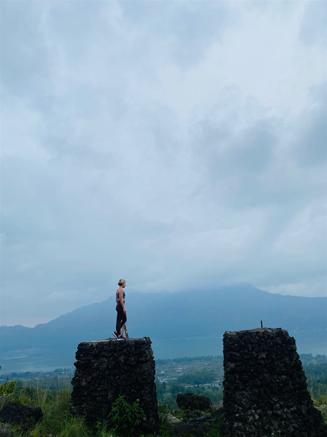 A figure standing on a stone ruin above a wide valley, a volcano ridge in the cloud behind