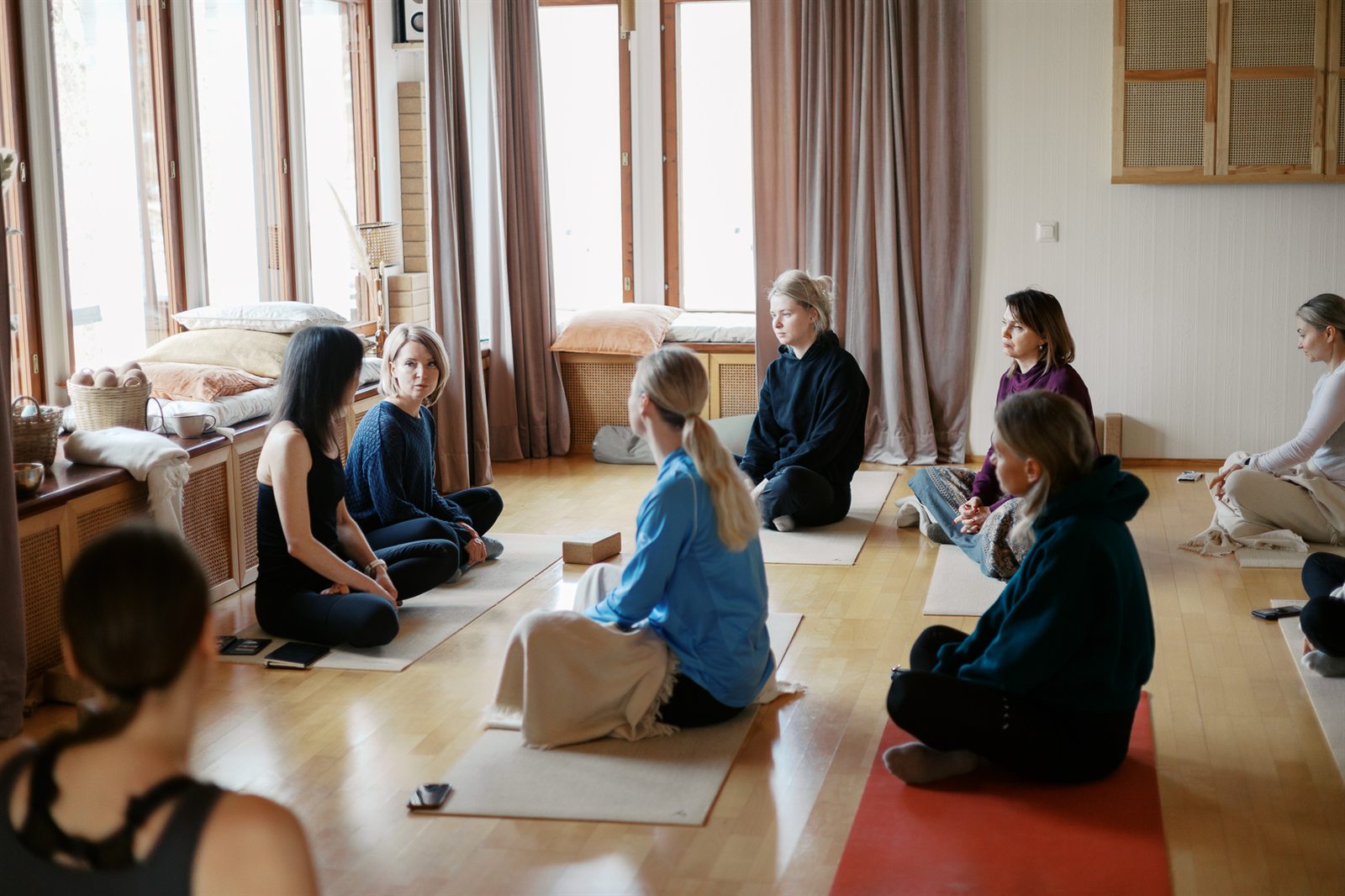 A circle of people sitting on mats in a bright wood-lined room, mid-conversation