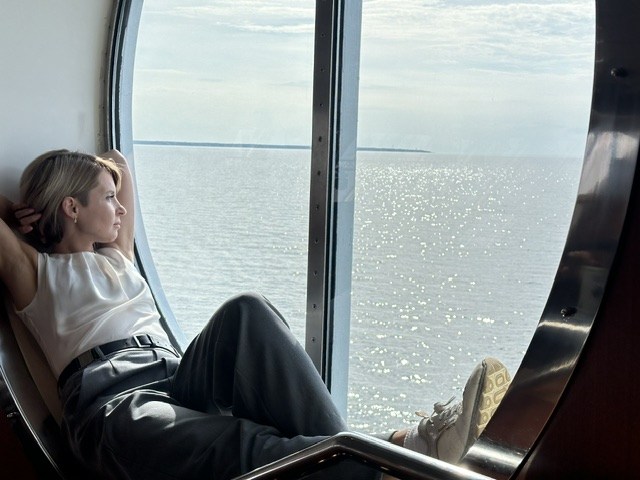Natasha resting in a round ship window, looking out over sunlit open sea